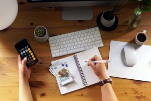 Um ambiente de trabalho organizado em uma mesa de madeira, onde uma pessoa está gerenciando suas finanças. A imagem mostra um notebook aberto com anotações de cálculos, dinheiro em espécie (notas e moedas de euro) e uma mão segurando um smartphone com a calculadora aberta. Também há um teclado sem fio, um mouse sobre um tapete, um monitor de computador, uma xícara de café e pequenos vasos com plantas.