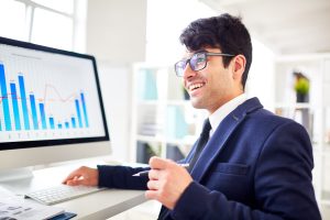 Um homem jovem, de terno azul e óculos, está sentado em um escritório moderno e bem iluminado, sorrindo enquanto analisa gráficos exibidos em um monitor de computador. Ele segura uma caneta e parece engajado em uma conversa ou apresentação.