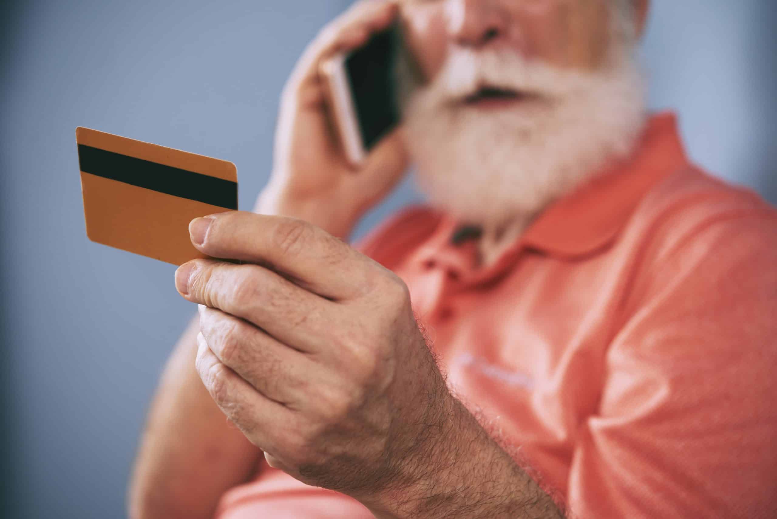Homem idoso de barba branca vestindo uma camisa polo laranja segura um cartão enquanto fala ao telefone, aparentando estar fazendo uma compra ou consulta financeira, possivelmente representando um momento de transação ou risco de golpe.