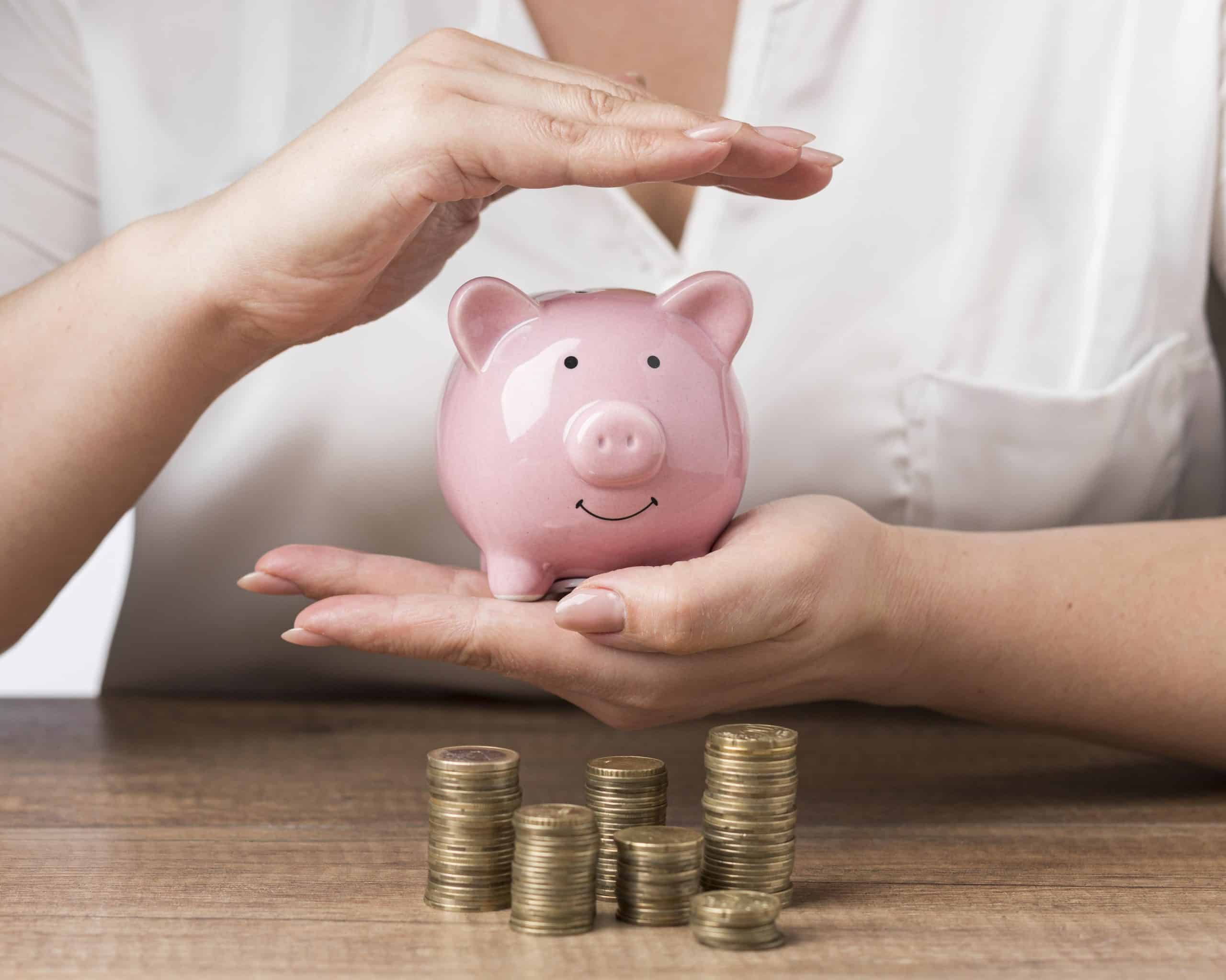 Mãos femininas segurando um cofrinho rosa em formato de porquinho sobre uma mesa de madeira, com pilhas de moedas douradas ao redor, simbolizando economia e planejamento financeiro.