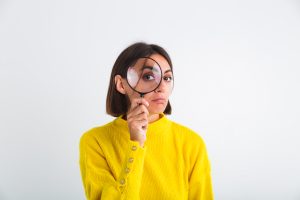 Uma mulher com um suéter amarelo segura uma lupa em frente ao rosto, ampliando um de seus olhos. Ela tem uma expressão curiosa e levemente séria, criando um efeito investigativo. O fundo branco e minimalista destaca sua figura e a cor vibrante da roupa.