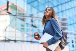 Uma jovem profissional caminha confiante em frente a um prédio moderno de vidro, segurando um laptop e um copo de café. Vestida com uma camisa azul e carregando uma mochila, ela transmite determinação e sucesso no ambiente corporativo. A imagem sugere a importância da ajuda de custo no trabalho, proporcionando benefícios que facilitam o dia a dia dos colaboradores, como transporte e alimentação.