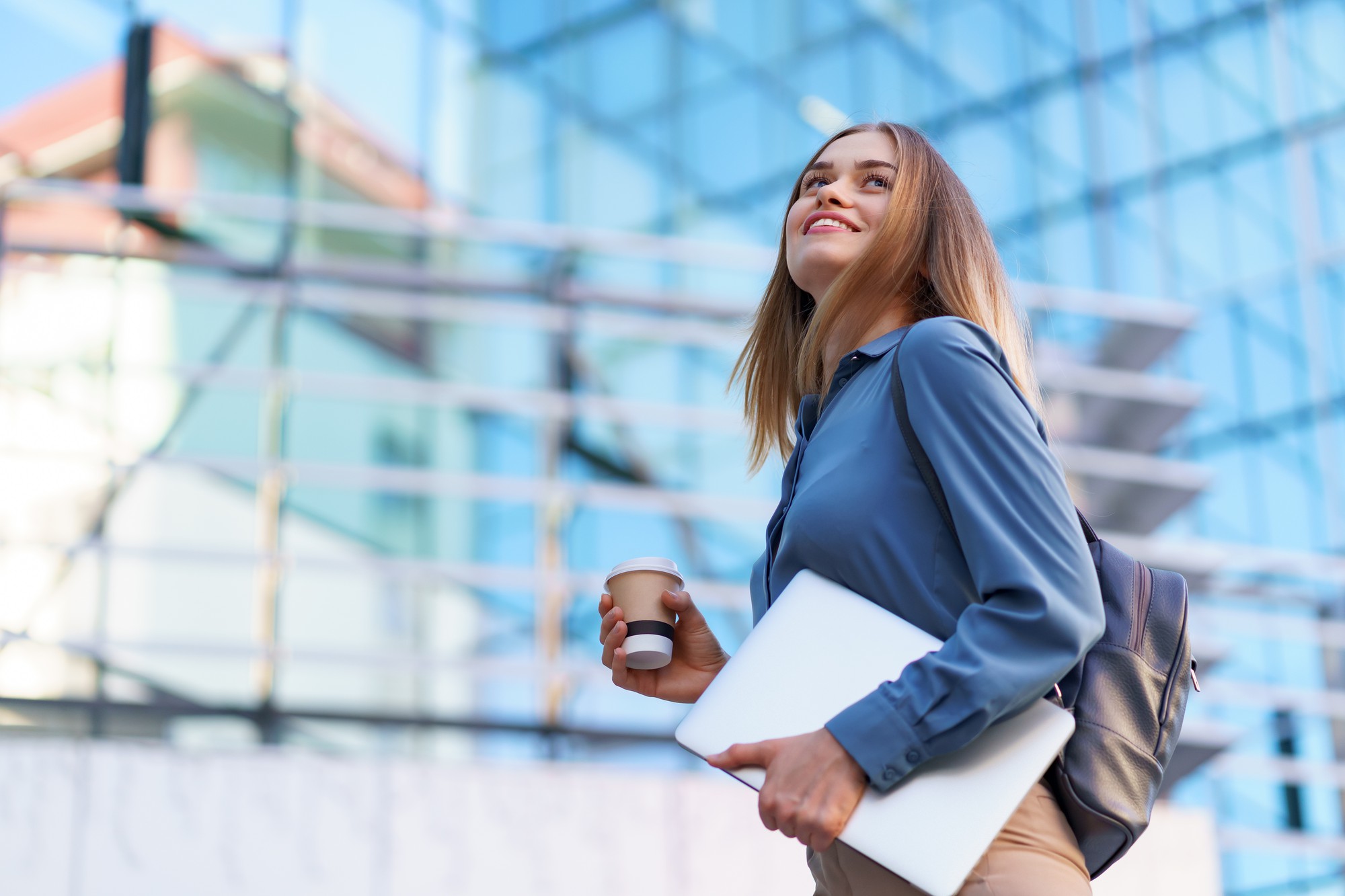 Uma jovem profissional caminha confiante em frente a um prédio moderno de vidro, segurando um laptop e um copo de café. Vestida com uma camisa azul e carregando uma mochila, ela transmite determinação e sucesso no ambiente corporativo. A imagem sugere a importância da ajuda de custo no trabalho, proporcionando benefícios que facilitam o dia a dia dos colaboradores, como transporte e alimentação.