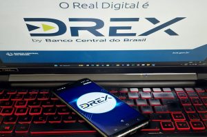 Um notebook com teclado iluminado exibe na tela o logotipo do Drex, a moeda digital do Banco Central do Brasil, acompanhado da frase "O Real Digital é Drex". Em primeiro plano, um smartphone também mostra o logotipo do Drex, reforçando a ideia de inovação e digitalização do sistema financeiro brasileiro. A imagem representa a modernização das transações financeiras e a implementação do real digital no país.