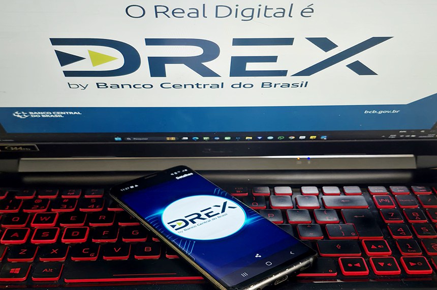 Um notebook com teclado iluminado exibe na tela o logotipo do Drex, a moeda digital do Banco Central do Brasil, acompanhado da frase "O Real Digital é Drex". Em primeiro plano, um smartphone também mostra o logotipo do Drex, reforçando a ideia de inovação e digitalização do sistema financeiro brasileiro. A imagem representa a modernização das transações financeiras e a implementação do real digital no país.