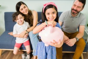 Menina sorridente segura um cofrinho rosa em formato de porquinho enquanto insere uma moeda, transmitindo a ideia de educação financeira infantil; ao fundo, seus pais e o irmão observam com orgulho e alegria, sentados em um sofá azul em um ambiente familiar e acolhedor.