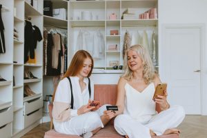 Duas mulheres, uma mais jovem de cabelo ruivo e outra mais velha de cabelo loiro, estão sentadas em um banco rosa dentro de um closet elegante. Ambas vestem roupas brancas sofisticadas e seguram smartphones, enquanto a mulher mais velha entrega um cartão de crédito preto para a mais jovem. O fundo exibe roupas, sapatos e acessórios organizados, sugerindo uma experiência de compras luxuosa, compras online ou conversas sobre finanças.