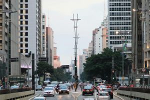 Imagem da movimentada Avenida Paulista em São Paulo no fim da tarde, com trânsito intenso nos dois sentidos, cercada por altos prédios comerciais e residenciais; ao fundo, o céu claro contrasta com as luzes dos edifícios e postes de iluminação modernos, enquanto pedestres se deslocam pelas calçadas e a estação de metrô Consolação aparece à esquerda.