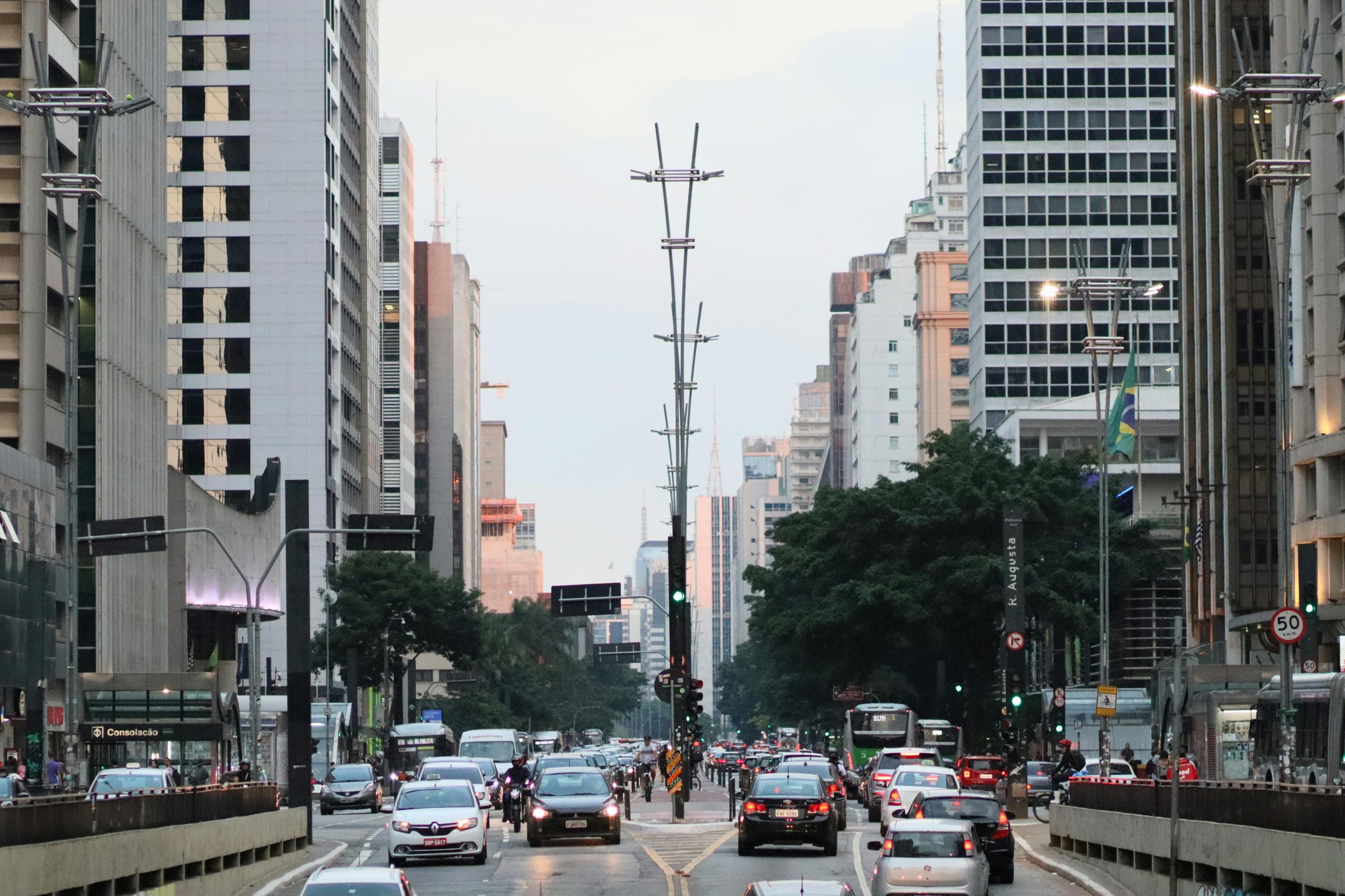 Imagem da movimentada Avenida Paulista em São Paulo no fim da tarde, com trânsito intenso nos dois sentidos, cercada por altos prédios comerciais e residenciais; ao fundo, o céu claro contrasta com as luzes dos edifícios e postes de iluminação modernos, enquanto pedestres se deslocam pelas calçadas e a estação de metrô Consolação aparece à esquerda.