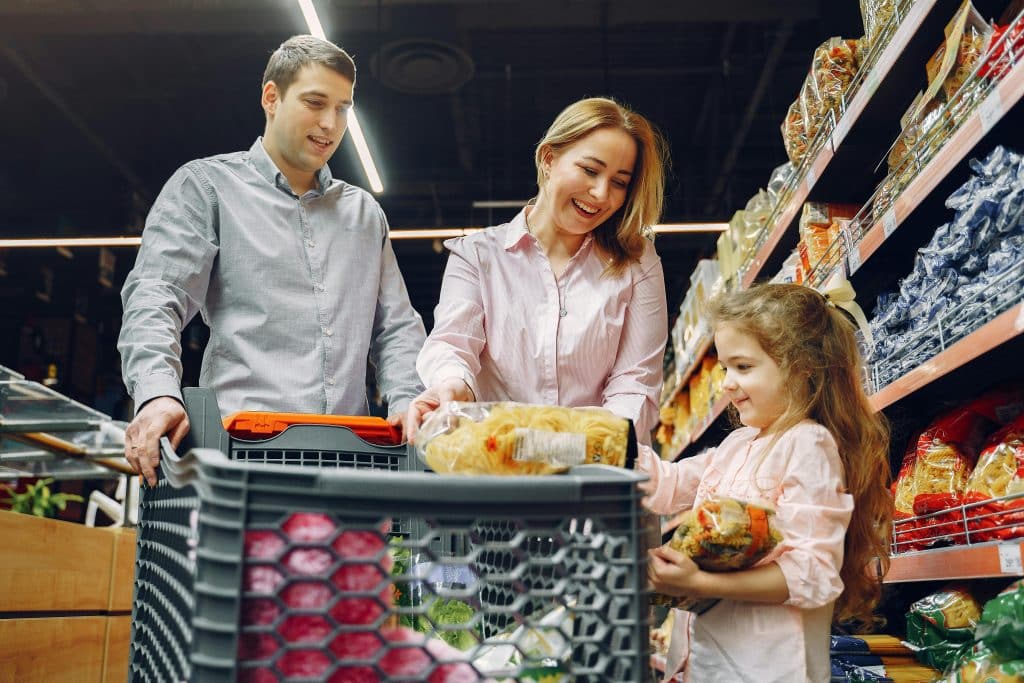 Família sorridente faz compras em um supermercado; o pai empurra o carrinho enquanto a mãe e a filha, que seguram pacotes de macarrão, escolhem produtos na prateleira de massas secas, demonstrando um momento de união e alegria no cotidiano.