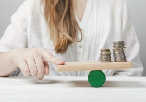 Uma mulher vestida de branco equilibra uma tábua de madeira sobre um cilindro verde, com pilhas de moedas dispostas em um dos lados. Ela toca suavemente a extremidade da tábua, representando o conceito de equilíbrio financeiro e gestão do dinheiro. A imagem simboliza a importância do planejamento para manter a estabilidade econômica e alcançar metas financeiras.