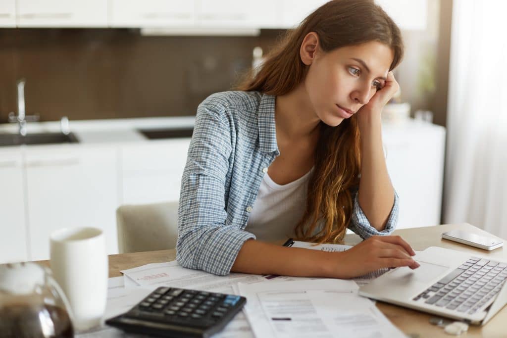 Mulher jovem com expressão preocupada, sentada à mesa de uma cozinha, cercada por contas, papéis e uma calculadora, enquanto olha para a tela do notebook. A imagem transmite o impacto da ansiedade financeira diante da organização das finanças pessoais.