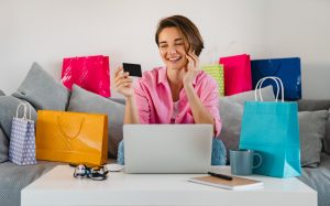 Mulher sorridente sentada em um sofá, cercada por sacolas de compras coloridas, segurando um cartão de crédito na mão enquanto usa um notebook. Ela parece estar fazendo compras online e falando ao celular, refletindo um momento de consumo ligado ao comportamento financeiro contemporâneo.
