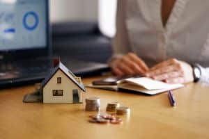 Pessoa analisando documentos financeiros ao lado de um notebook, com uma maquete de casa e pilhas de moedas sobre a mesa, simbolizando planejamento e proteção patrimonial por meio do seguro prestamista, que garante a quitação de dívidas em situações imprevistas.