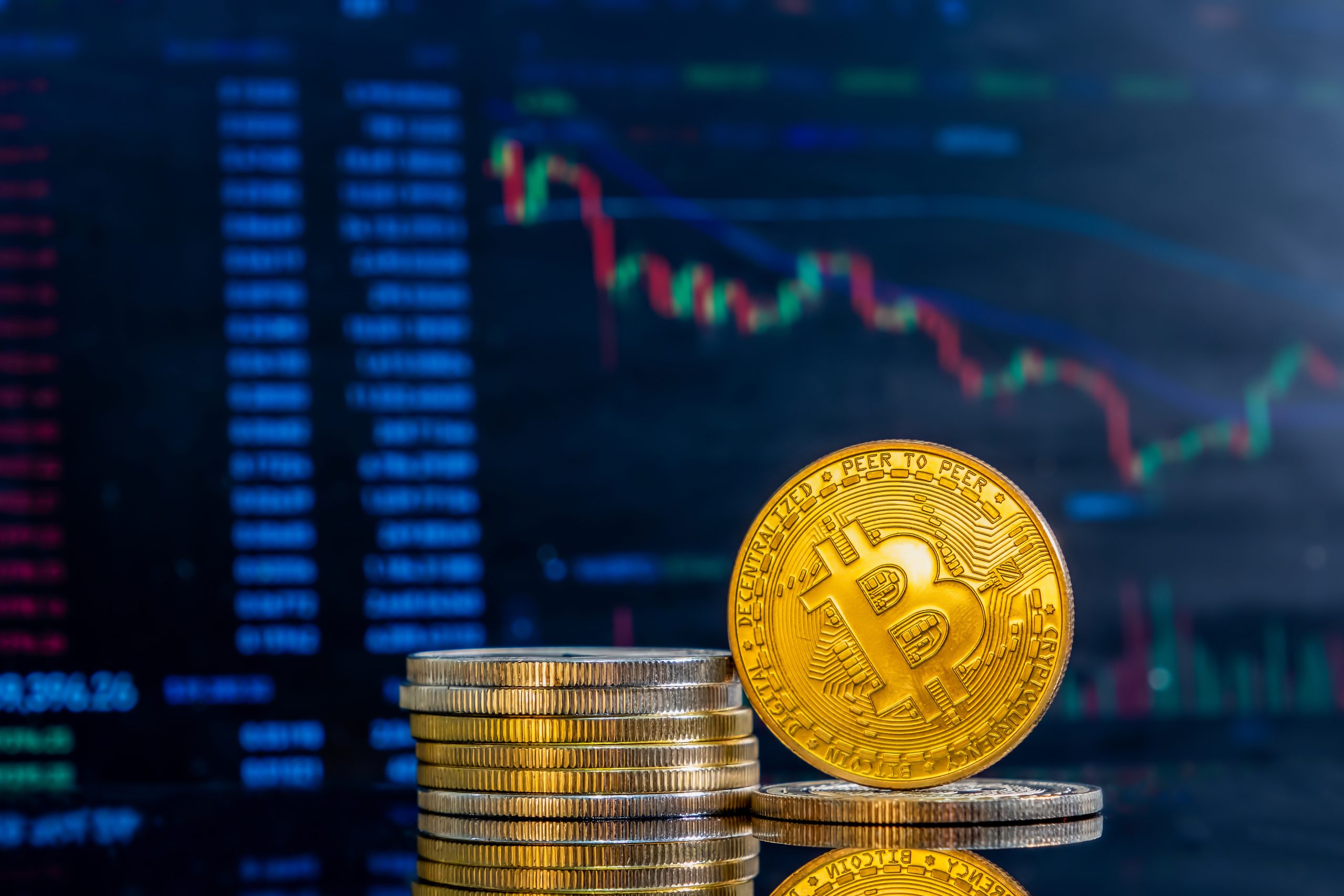 Pilha de moedas físicas representando criptomoedas, com destaque para uma moeda dourada de Bitcoin em primeiro plano, e ao fundo uma tela com gráficos financeiros e cotações de mercado digital.