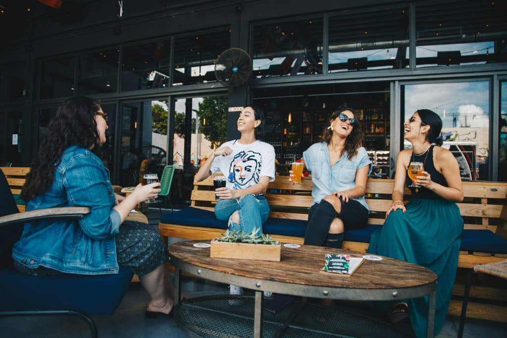 Quatro mulheres jovens sorrindo e conversando animadamente em um bar ou café ao ar livre, cada uma segurando uma bebida, transmitindo um momento de descontração e amizade.