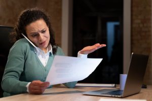 Mulher ao telefone com expressão de frustração enquanto analisa um documento, sentada em frente ao notebook, ilustrando os possíveis riscos de emprestar sua conta e seus documentos, como transtornos com cobranças indevidas ou envolvimento em fraudes.