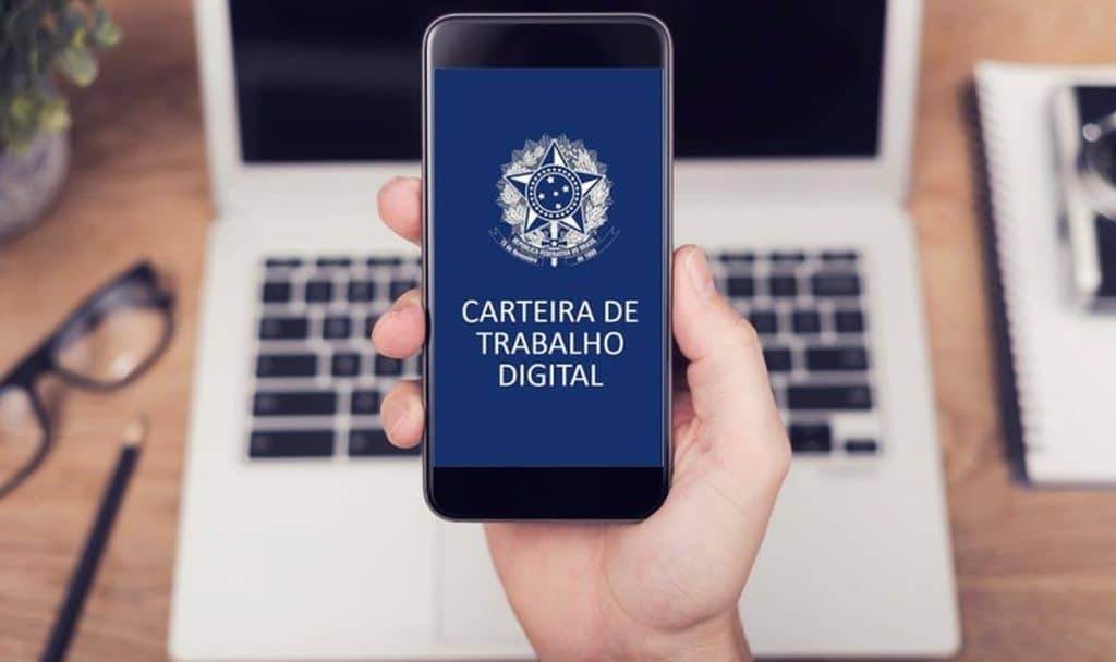 Pessoa segura um smartphone exibindo a tela inicial da CTPS digital, com o emblema da República Federativa do Brasil, em um ambiente de trabalho com notebook e caderno ao fundo.