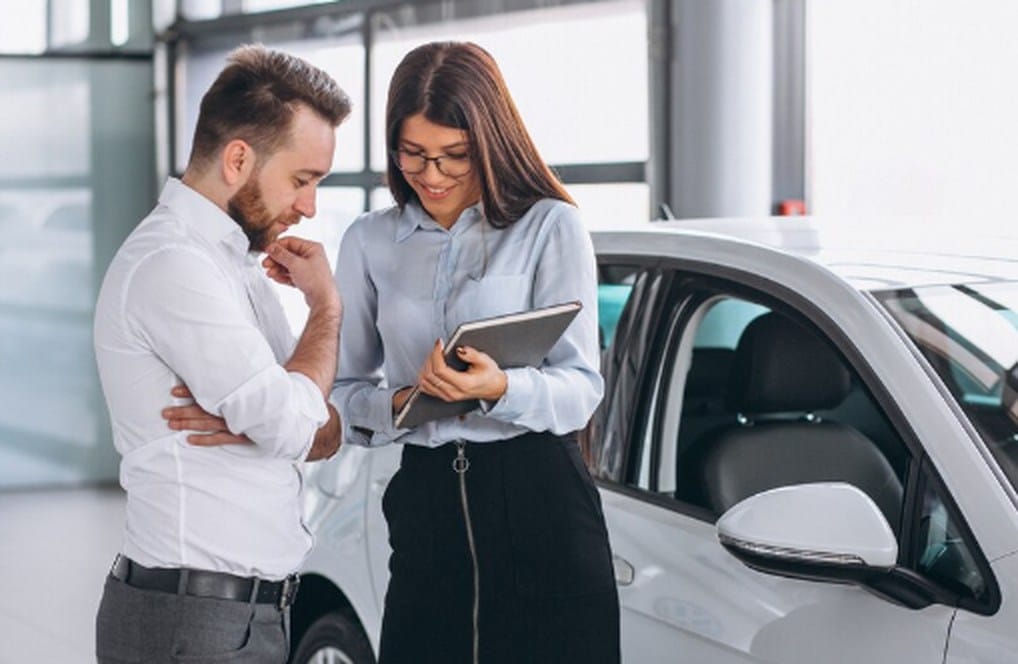 Consultora explica opções de consórcio ou financiamento para a compra de um veículo a um cliente interessado, enquanto ambos analisam informações em um tablet ao lado de um carro dentro de uma concessionária moderna e bem iluminada.