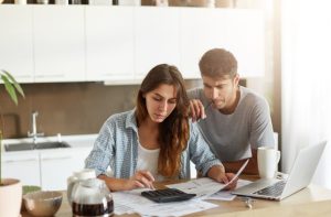 Casal analisa documentos e faz cálculos com uma calculadora e notebook sobre a mesa, representando o planejamento financeiro para refinanciamento de empréstimo em ambiente doméstico.