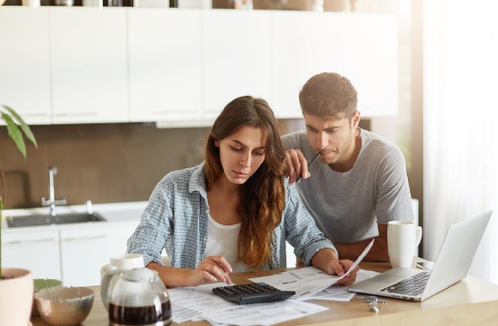 Casal analisa documentos e faz cálculos com uma calculadora e notebook sobre a mesa, representando o planejamento financeiro para refinanciamento de empréstimo em ambiente doméstico.