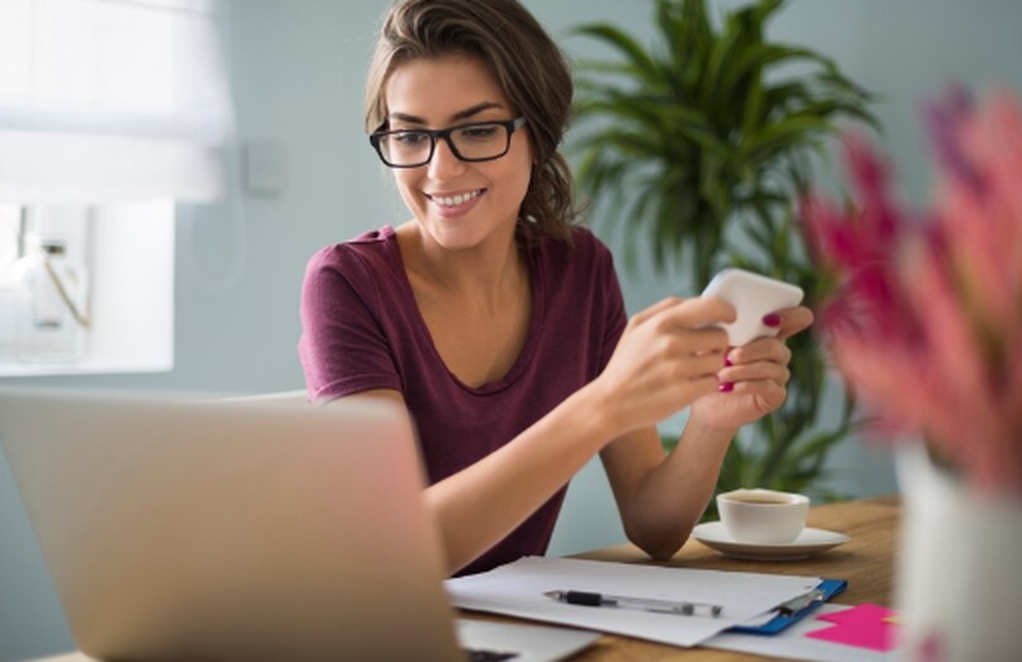Mulher sorridente trabalha em casa com notebook e celular, analisando anotações sobre a mesa, representando organização e busca por oportunidades de renda extra.