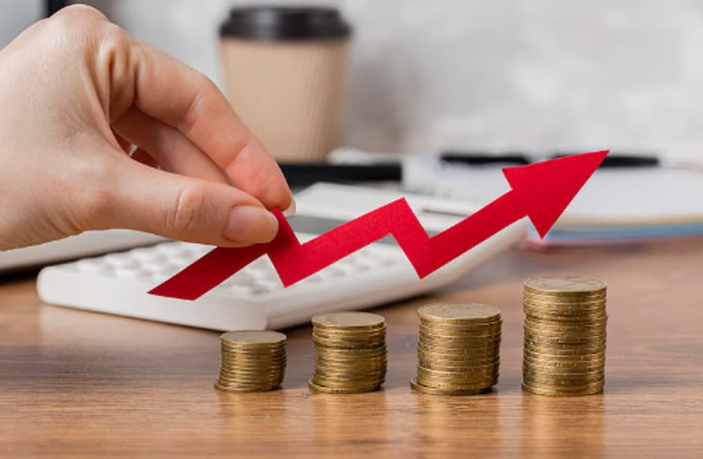 Mão segurando uma seta vermelha apontando para cima sobre pilhas crescentes de moedas, simbolizando o aumento do IOF e o impacto do imposto sobre operações financeiras.