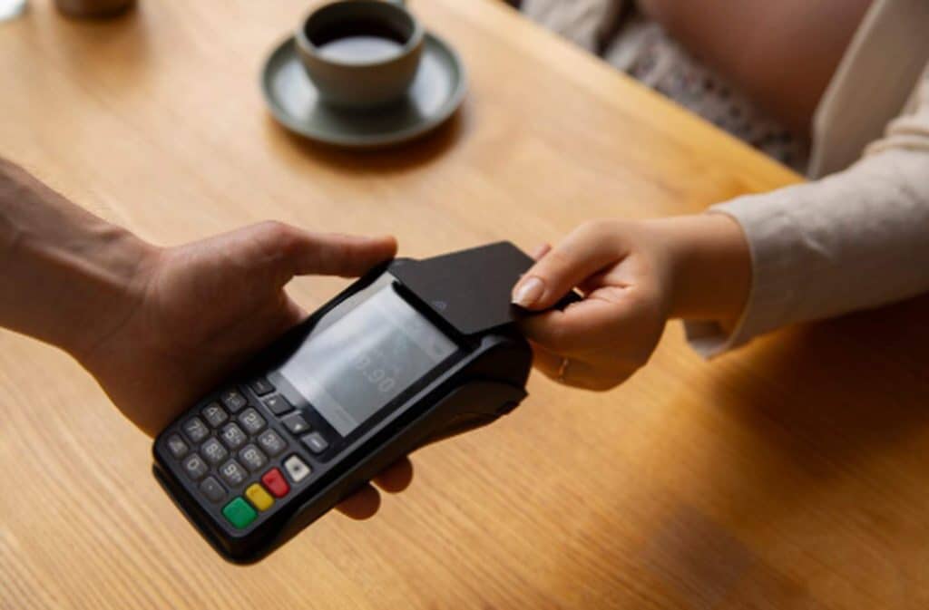 Pessoa realizando um pagamento por aproximação com cartão em uma maquininha de cartão sobre a mesa de madeira, representando praticidade e tecnologia nas transações financeiras.