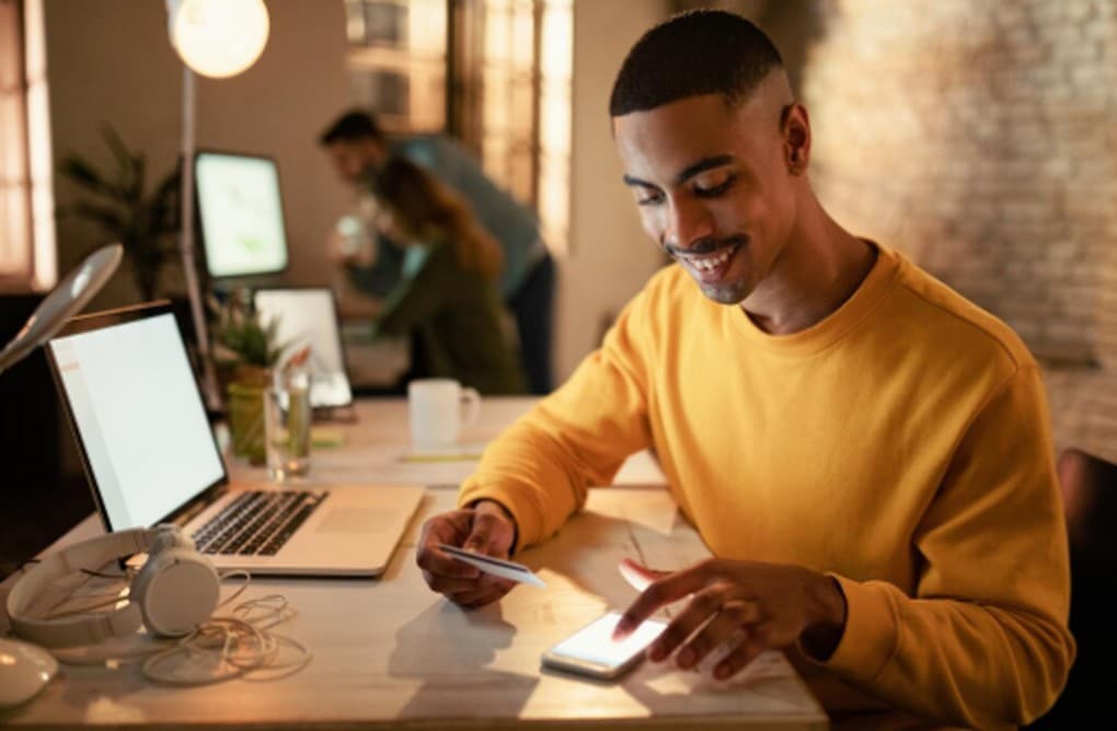 Homem sorridente usando smartphone e notebook em um escritório moderno, representando a praticidade e facilidade de solicitar crédito pré-aprovado de forma online e segura.