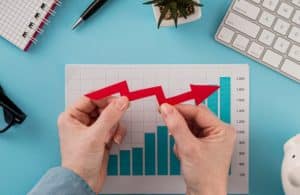 Mãos segurando uma seta vermelha apontando para cima sobre gráfico de barras azuis em um escritório, representando o impacto da inflação no crescimento dos preços e no poder de compra.