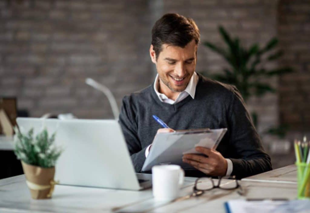 Homem sorridente sentado em seu escritório, analisando documentos ao lado do notebook, representando organização financeira e as oportunidades oferecidas pelo microcrédito produtivo para pequenos empreendedores.