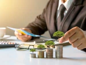 Homem de negócios organizando documentos financeiros em uma mesa, ao lado de pilhas de moedas que crescem progressivamente e sustentam pequenas árvores, simbolizando crescimento sustentável, planejamento e a importância do autocuidado e saúde financeira.