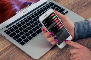 Pessoa segurando um smartphone com gráficos e cotações exibidos na tela, em frente a um notebook, representando o uso de tecnologia para análise de investimentos e controle financeiro por app.