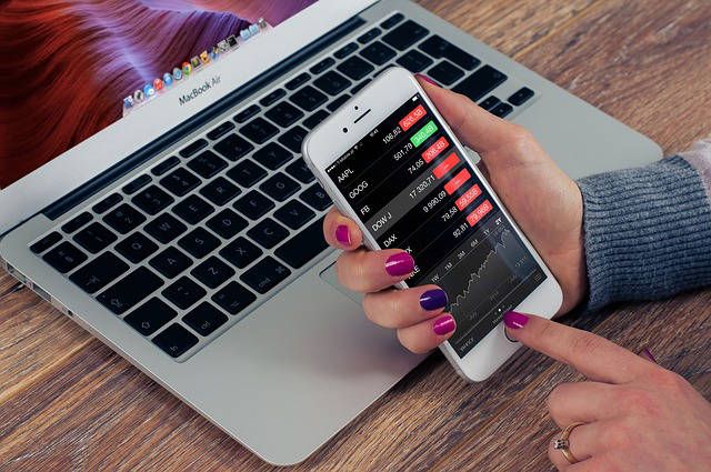 Pessoa segurando um smartphone com gráficos e cotações exibidos na tela, em frente a um notebook, representando o uso de tecnologia para análise de investimentos e controle financeiro por app.
