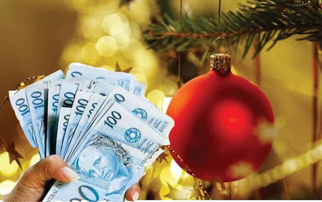 Mão segurando várias cédulas de 100 reais ao lado de uma bola vermelha de Natal em árvore decorada, simbolizando o impacto das despesas sazonais no orçamento durante as festas de fim de ano.