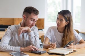 Casal jovem discutindo enquanto analisa números em uma calculadora e anotações, representando como as finanças podem impactar o seu relacionamento no dia a dia.