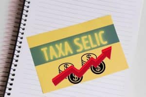 Cartão amarelo com a frase “Taxa Selic” e uma seta vermelha apontando para cima sobre fundo de caderno, simbolizando o aumento da Selic e seus impactos nas finanças e nos investimentos.
